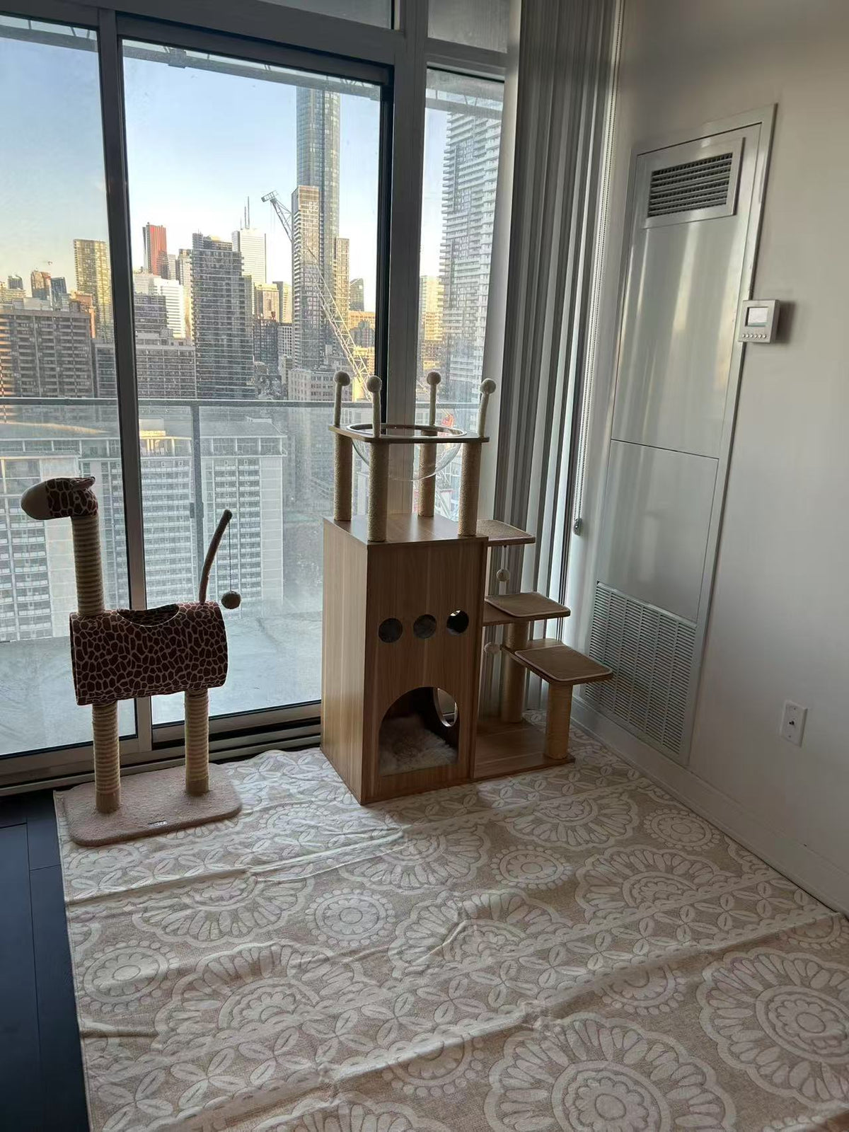 A modern cat tree and cat perch set in a bright, sunlit room with a city skyline view. The set includes a tall wooden tower with multiple levels, a giraffe-style perch with a leopard-print cushion, and a patterned floor mat.